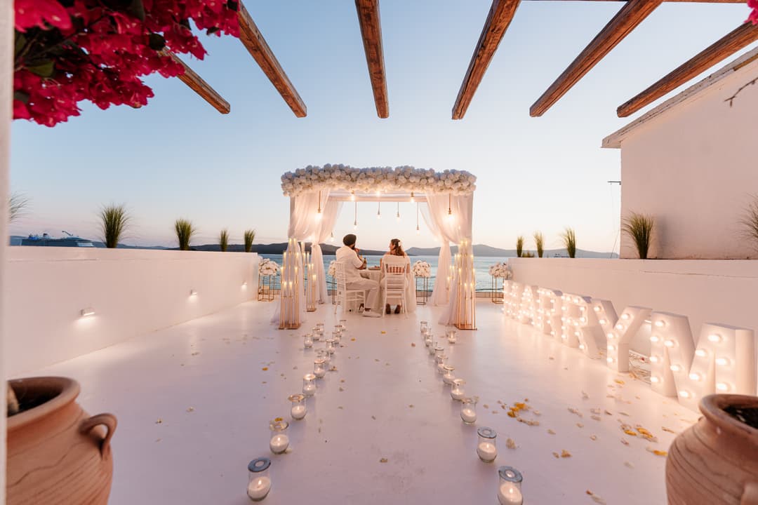 A couple at a small dinner table under the lit white canopy at AION at dusk, candles along the terrace and marquee letters glowing beside them