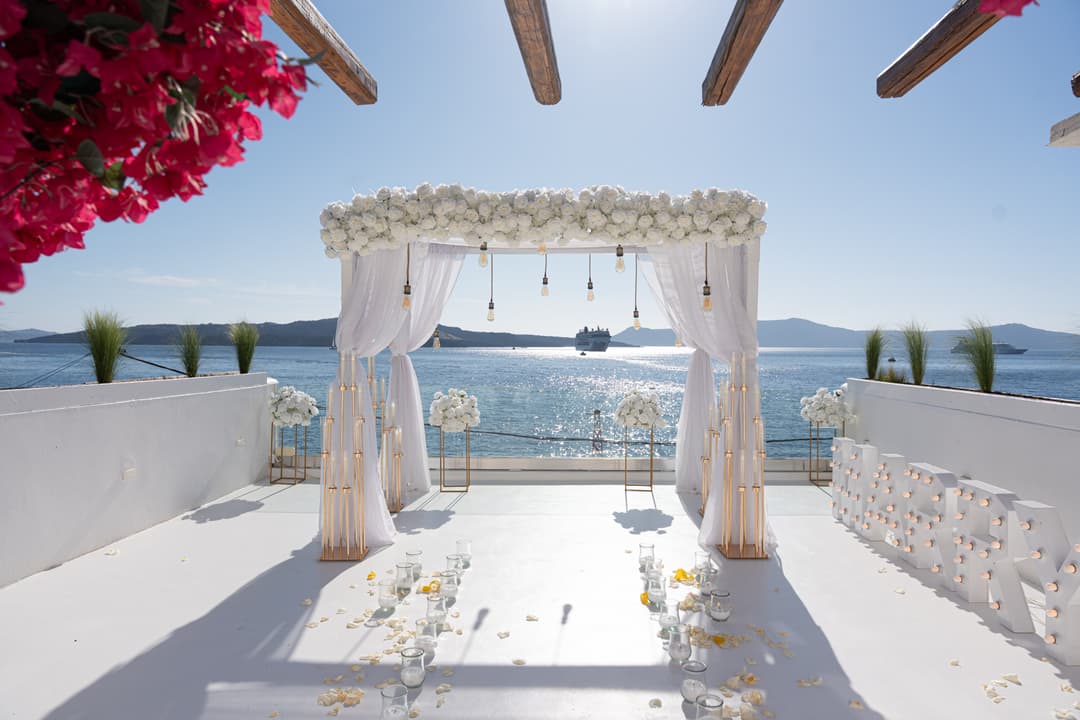 The white set at AION: a white-rose canopy over open water under a bougainvillea pergola, candle stands flanking the aisle, marquee letters at the rail