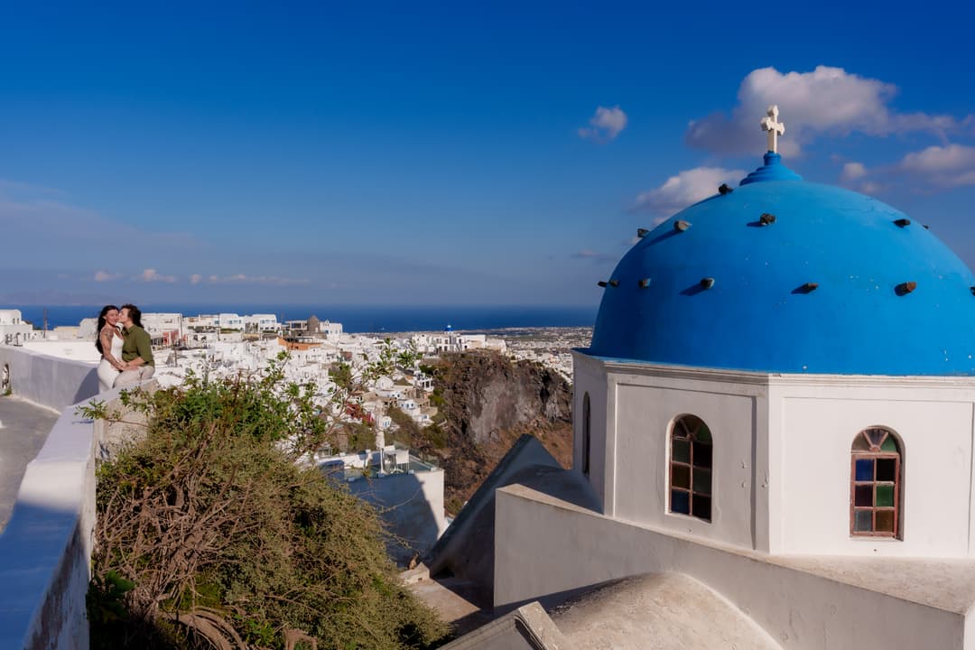 A blue-domed church with its white cross in the foreground, the village and open sea falling away behind it