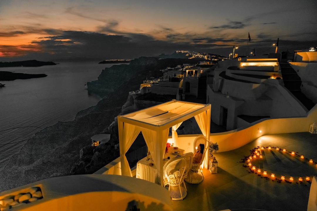 A dinner canopy glowing after dark on the caldera rim, village lights spilling down the cliff