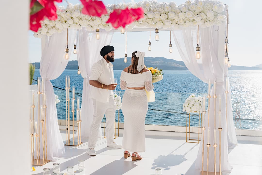 A couple under a white-rose canopy hung with filament lamps, the open sea and the caldera rim behind them