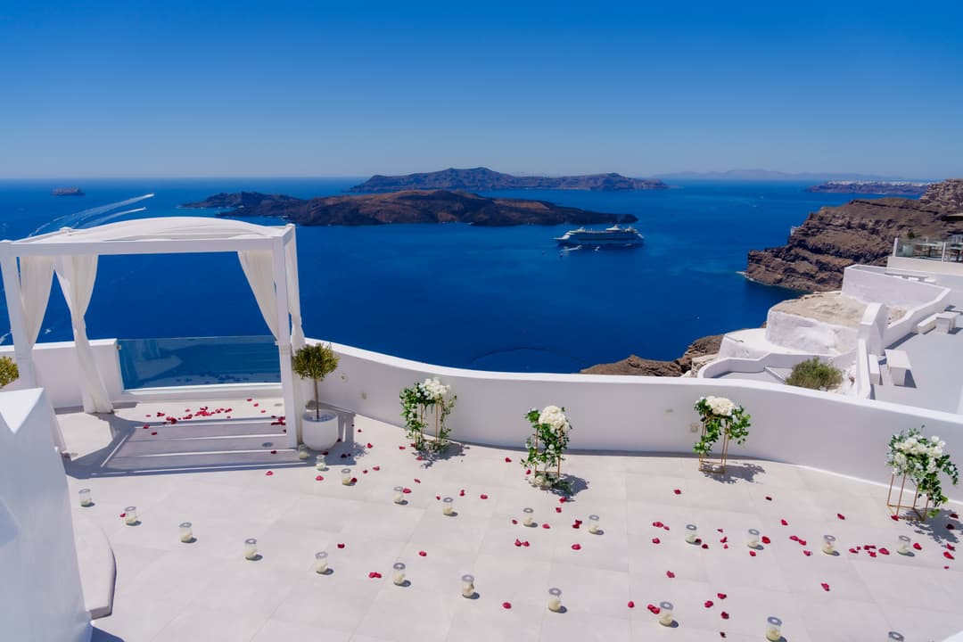 A caldera terrace fully dressed and waiting: white canopy, flower stands, candles and rose petals, the sea far below