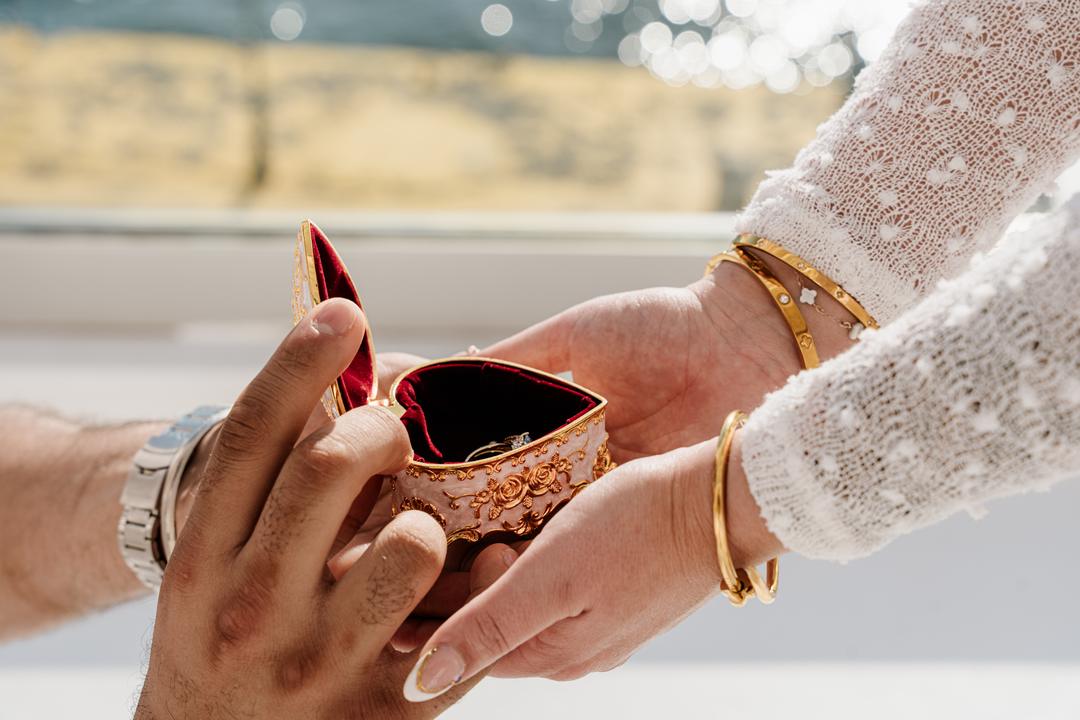 A gold filigree ring box passed between two hands, a solitaire ring inside on red velvet