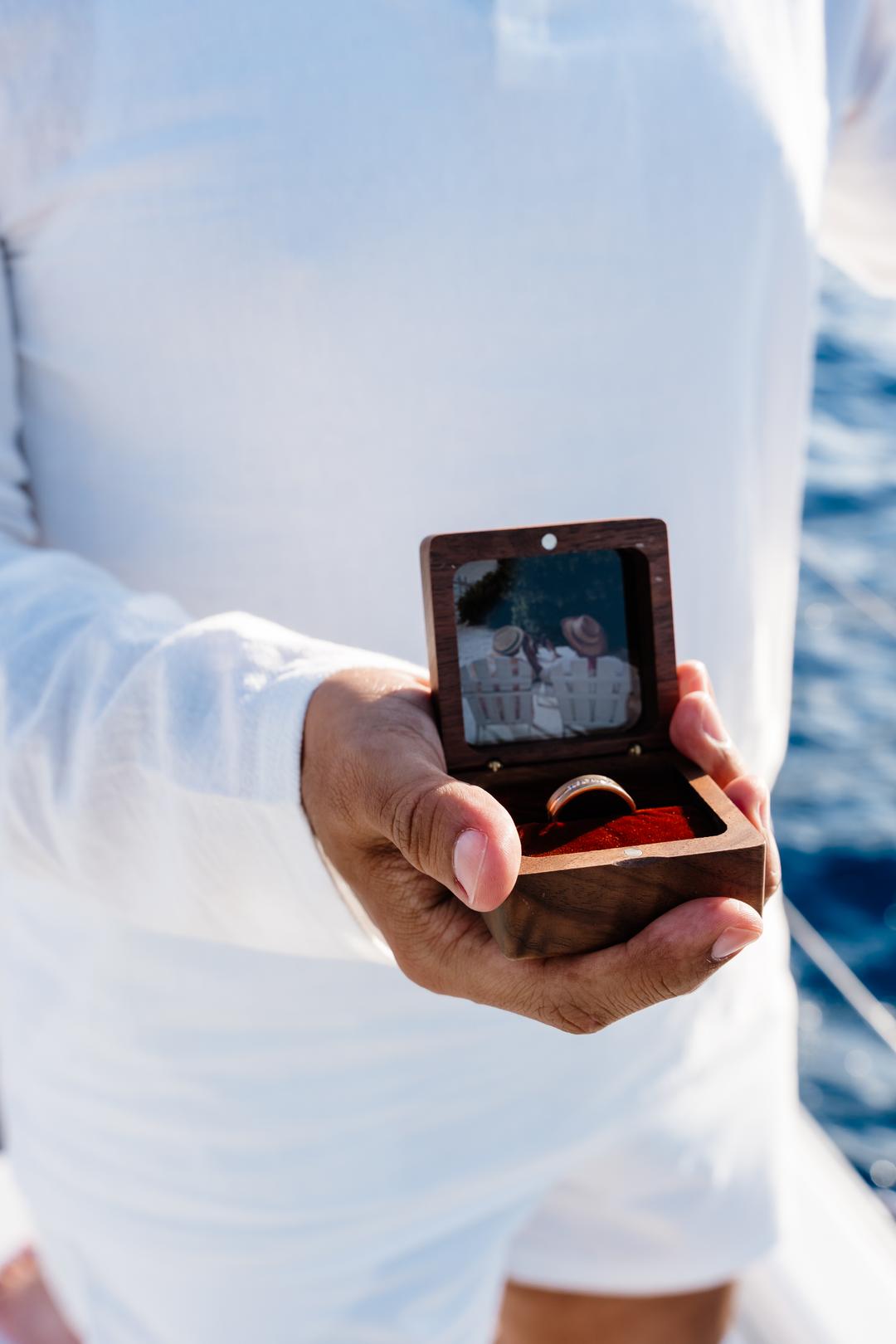 A hand holding a wooden ring box open behind his back, a small photograph mounted inside the lid