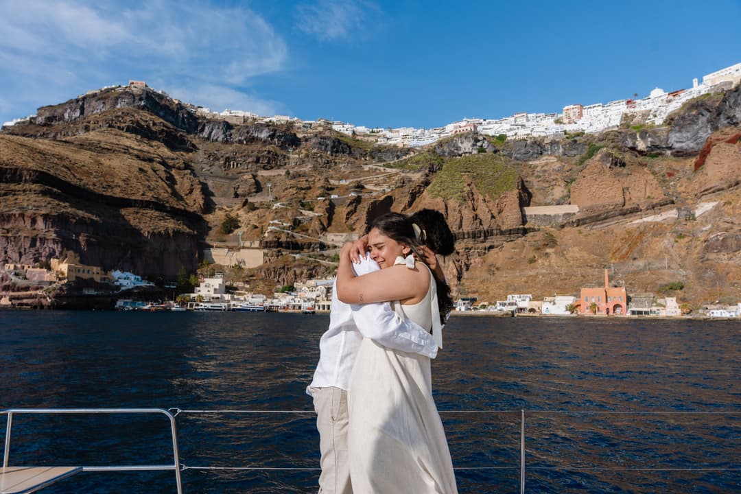 An embrace on deck below the Old Port of Fira: the zig-zag path and cable car climbing the cliff, the white town along the rim high above