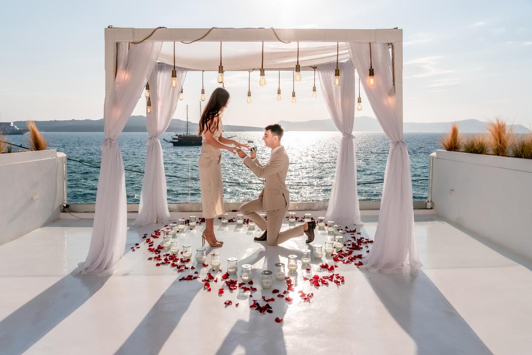 Kneeling beneath a white canopy at a waterfront venue, petals across the deck