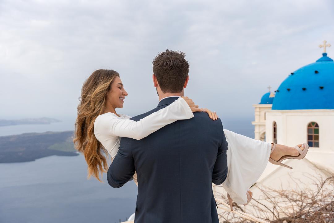 A man carrying a woman in his arms on a terrace, a blue dome and the flat gray water of the caldera behind them