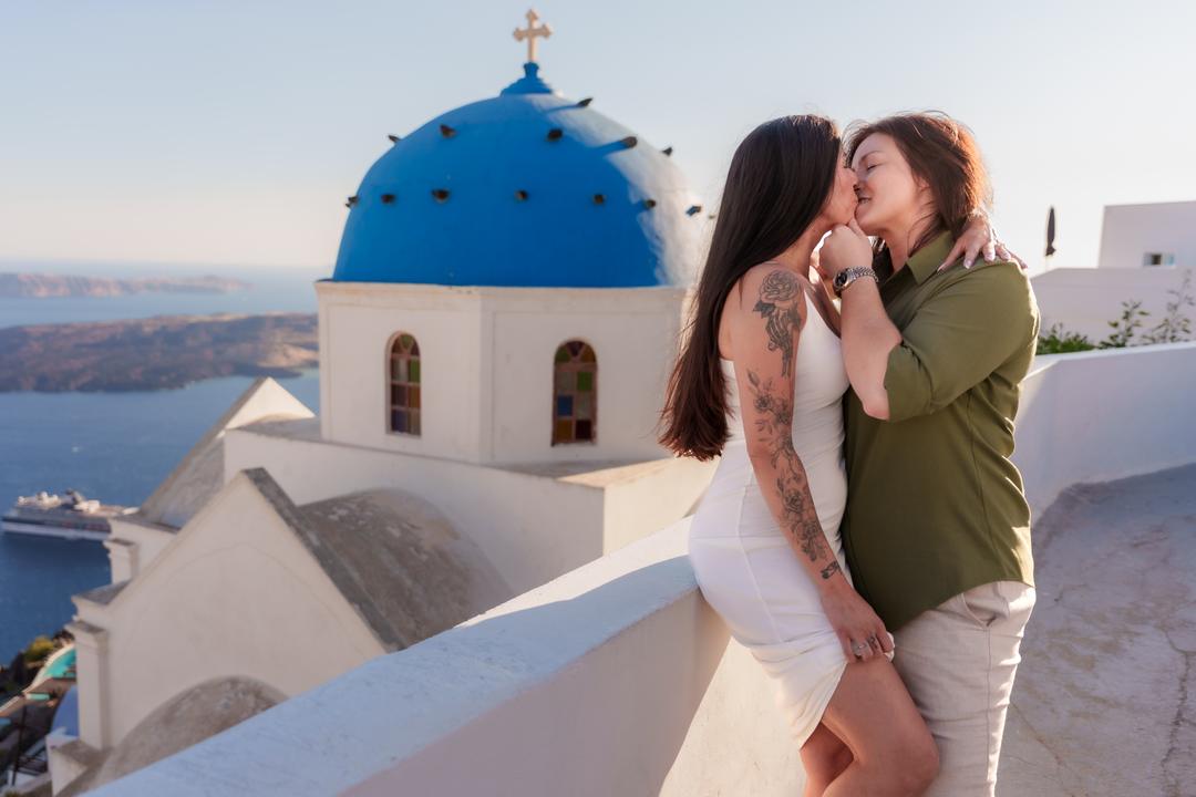 A couple kissing on a white terrace beside a blue-domed church, the caldera water flat behind them