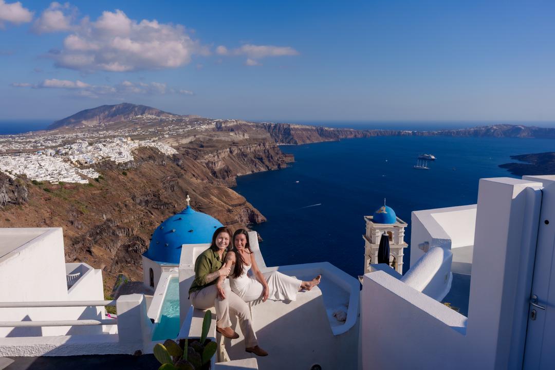 A couple sitting on a white wall above a blue dome and a bell tower, the caldera and the far rim opening out behind them