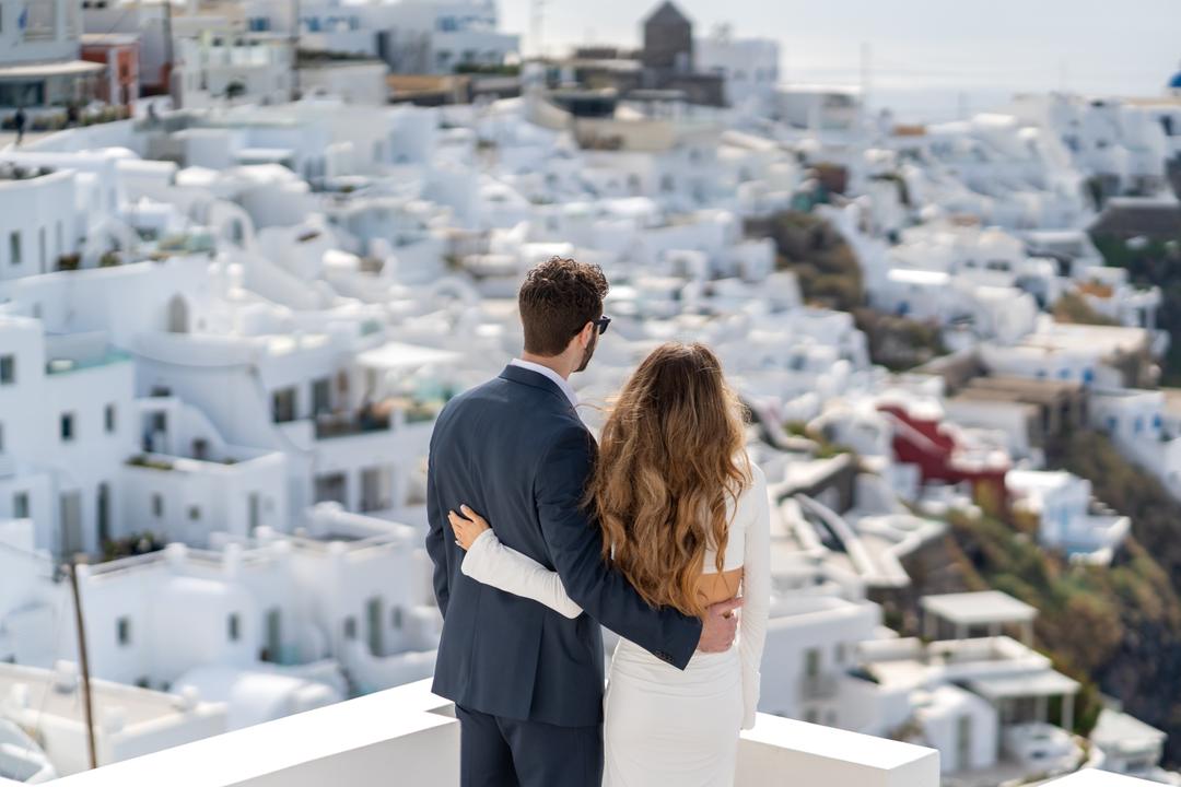 A couple with their backs turned at a white wall, looking out over the whitewashed rooftops of the village below