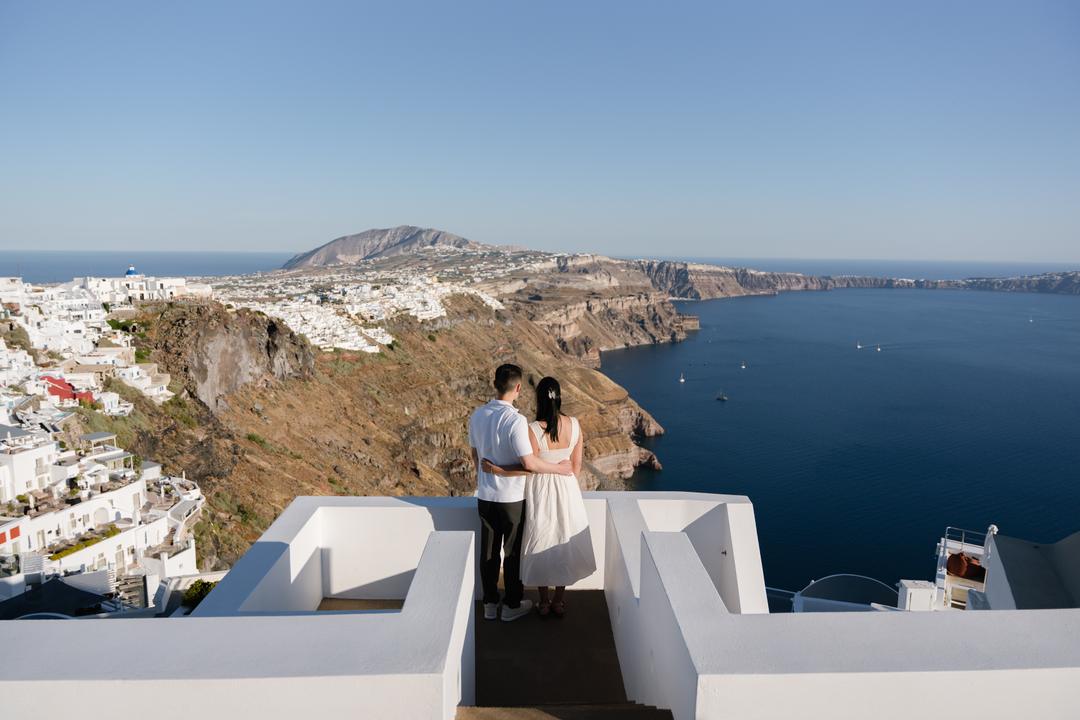 A couple standing at the end of a white terrace with the whole caldera, the stacked village and the deep blue water spread out behind them