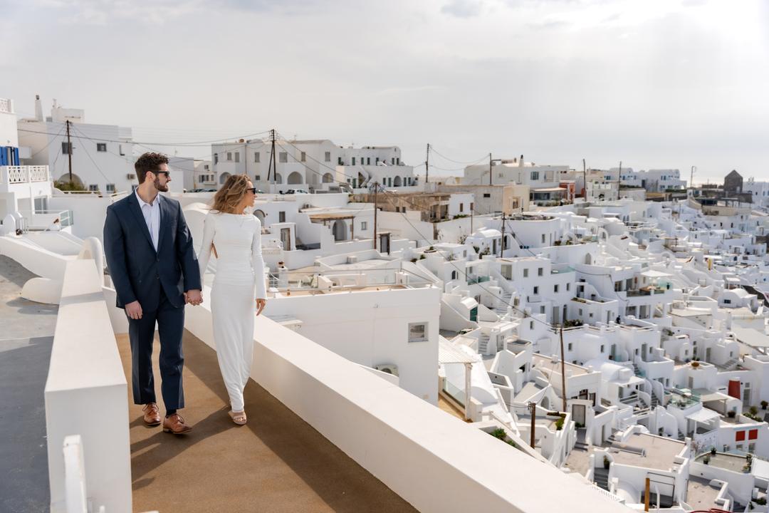 A couple walking a long white terrace above the stacked white roofs of the village