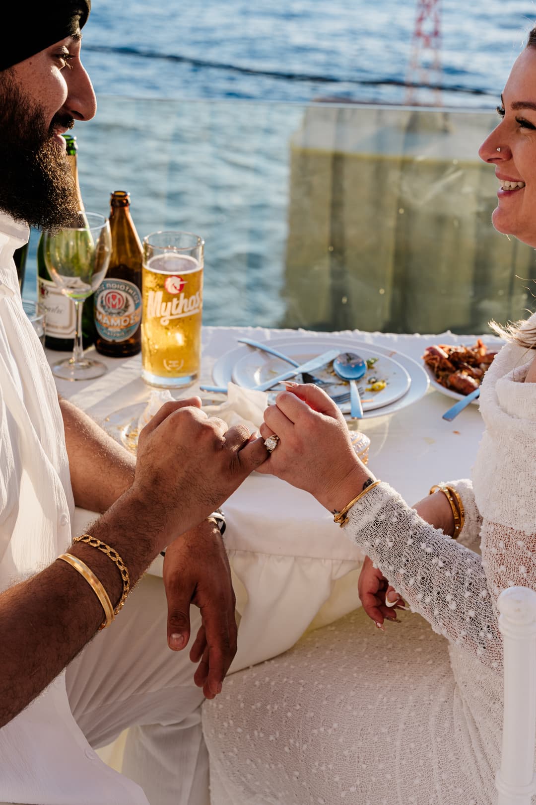 Two linked hands across a finished dinner table by the water, rings catching the late sun