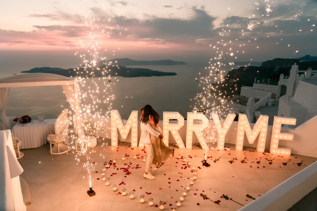 Cold-spark fountains lighting Marry Me letters on a caldera terrace at dusk