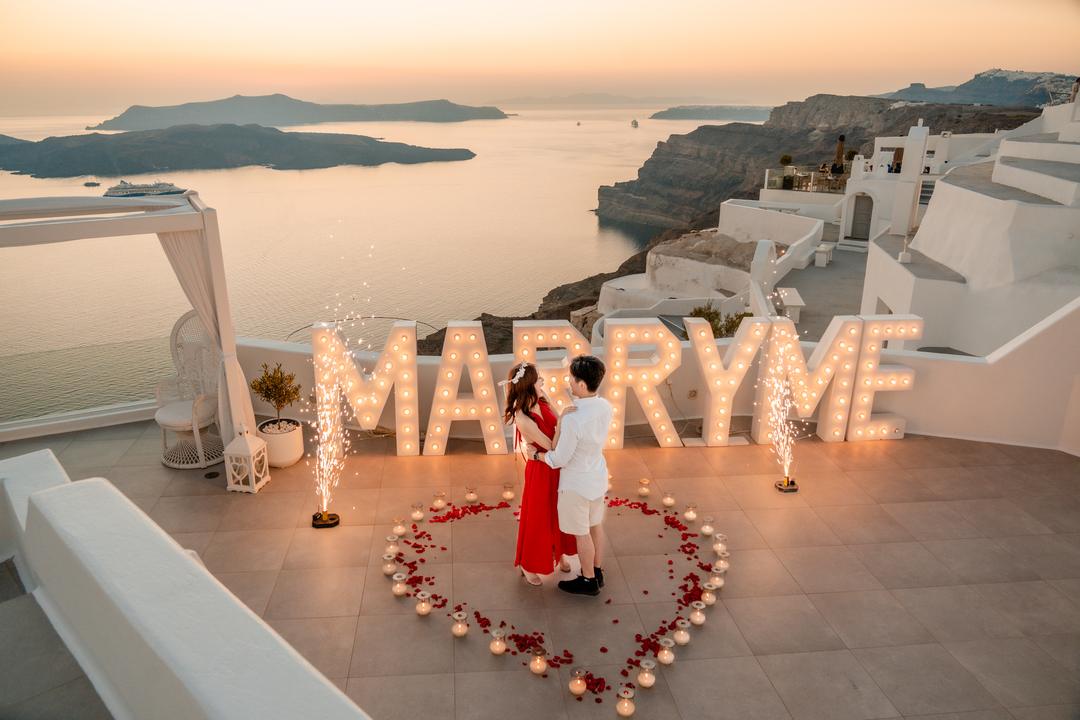 Marry Me letters glowing beside a petal heart on a caldera terrace at dusk