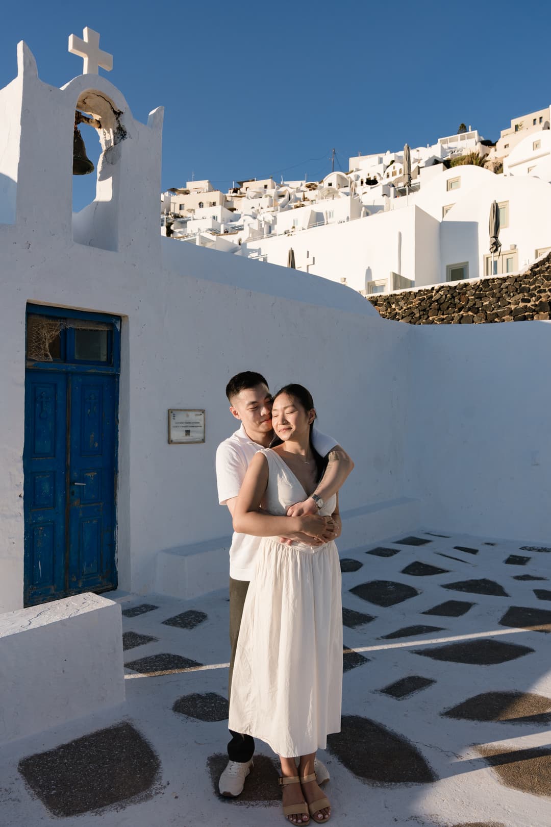 A couple standing quietly in a whitewashed parish courtyard, the chapel's bell and blue door beside them and the village climbing behind