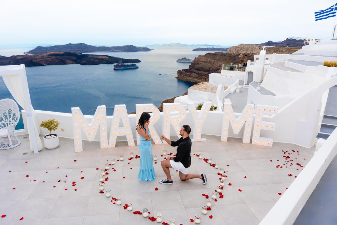 A real proposal mid-question: kneeling beside Marry Me letters and rose petals on a white caldera terrace