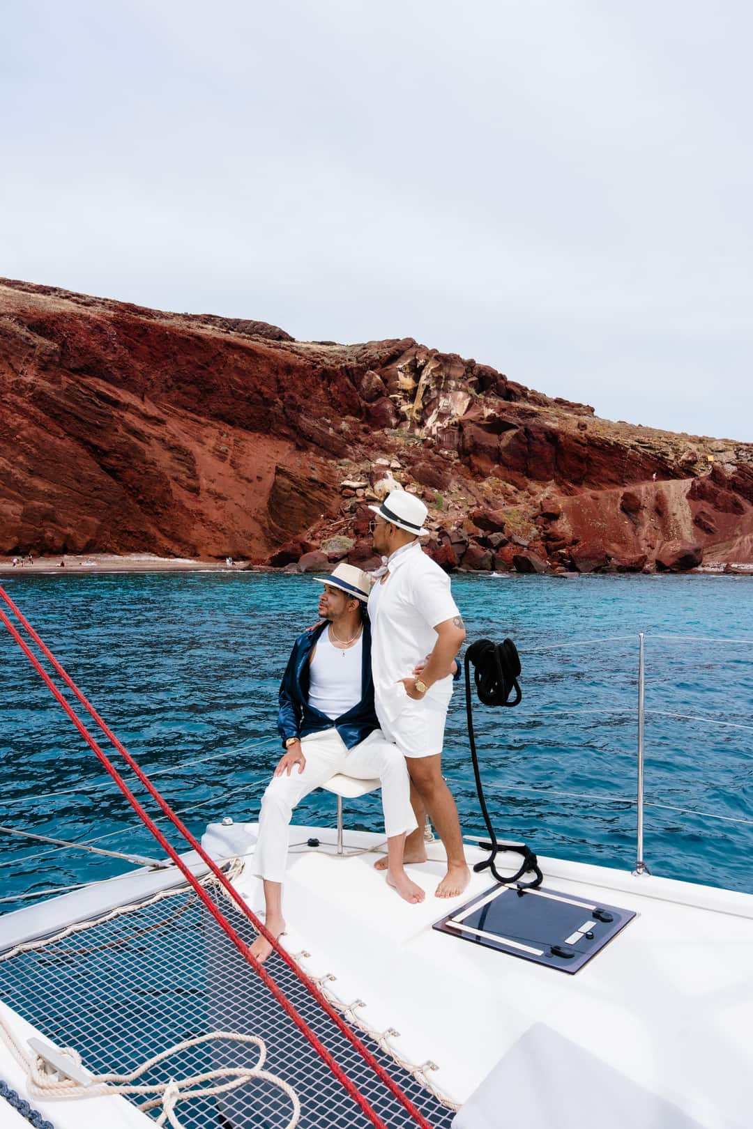 Red Beach from the water: the crimson cliff amphitheatre rising over the sand, a couple watching from the bow of a catamaran