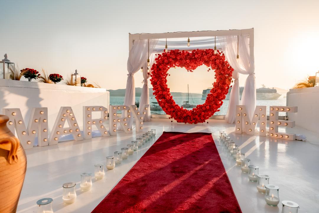 A red-rose heart arch, Marry Me letters and candles styled on a caldera terrace at dusk