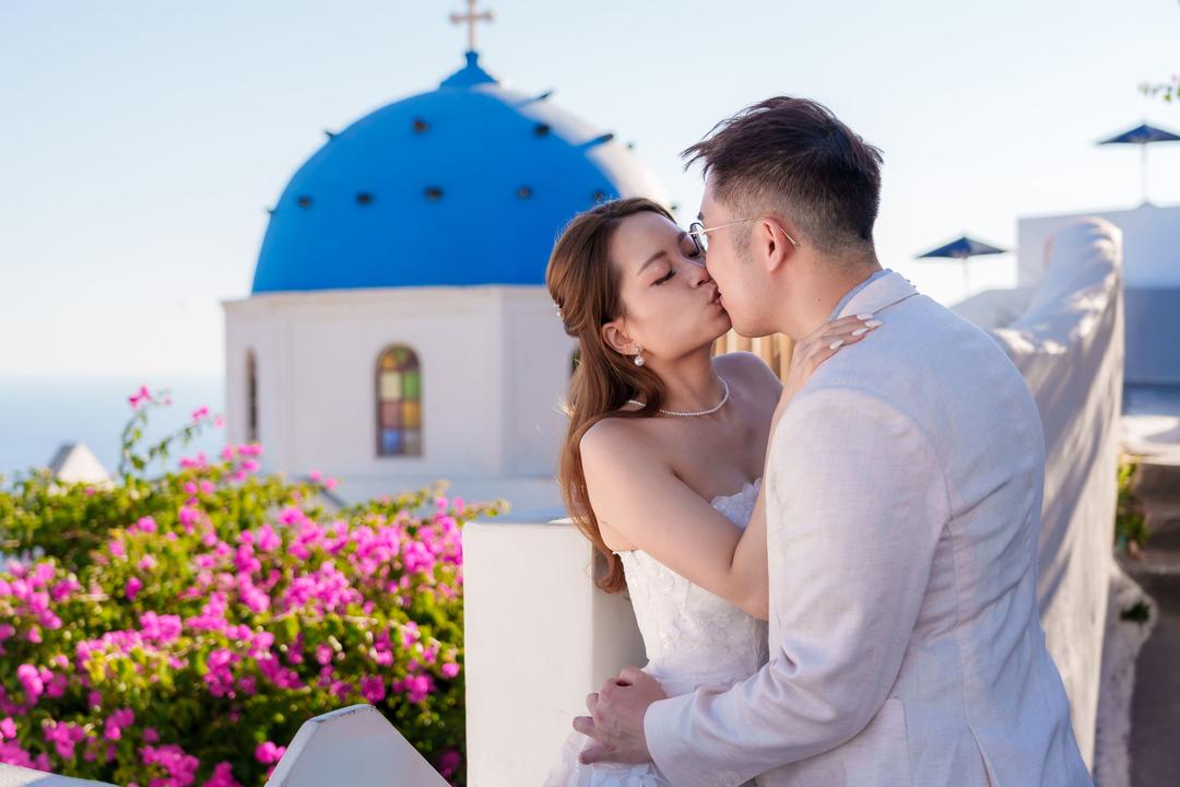 A couple kissing beside a blue-domed church with magenta bougainvillea in flower below them