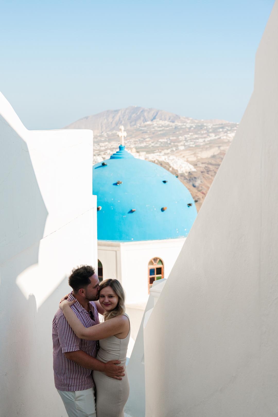 A couple embracing in a gap between two white walls, a single blue church dome and the village beyond them
