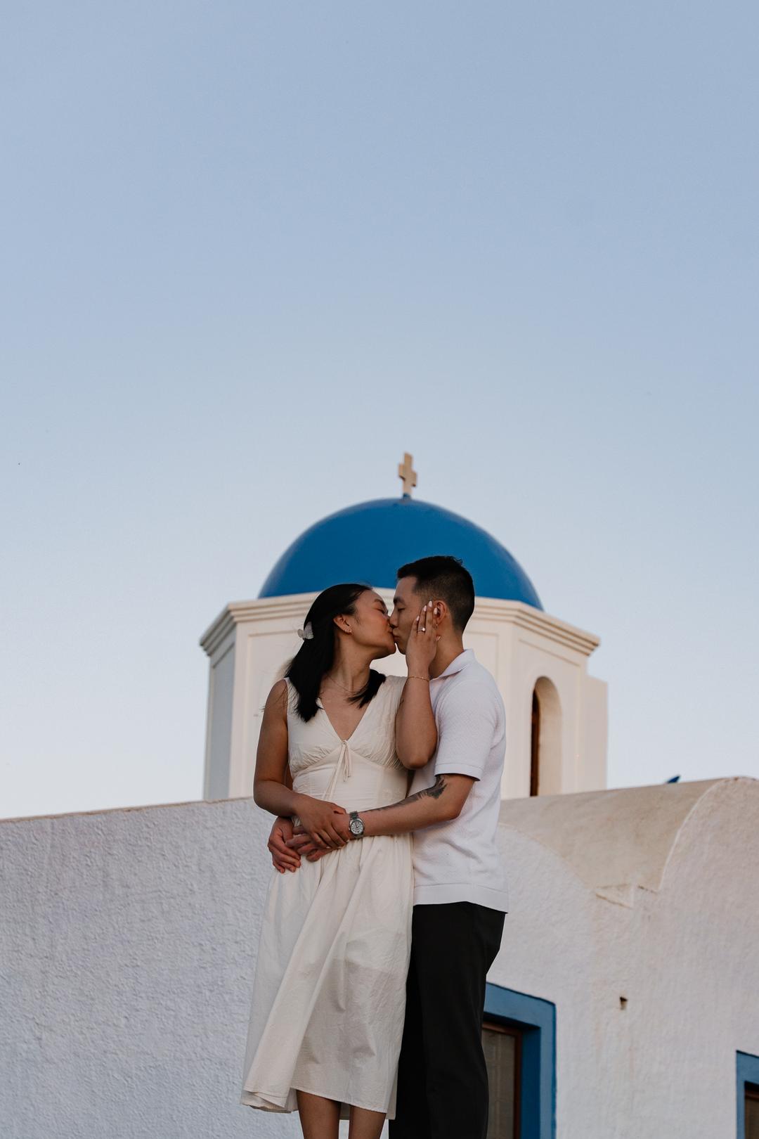 A couple kissing in front of a blue-domed chapel under a clear sky