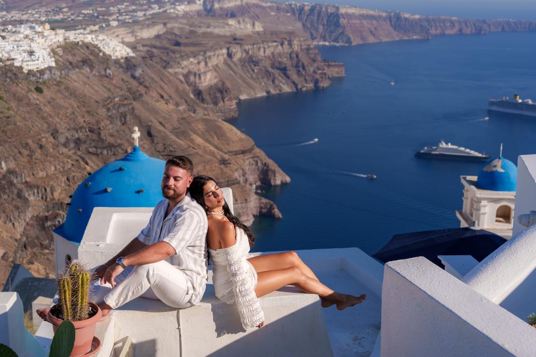 A couple sitting on a whitewashed ledge above a blue church dome, the full caldera, the cliff villages and anchored yachts behind them