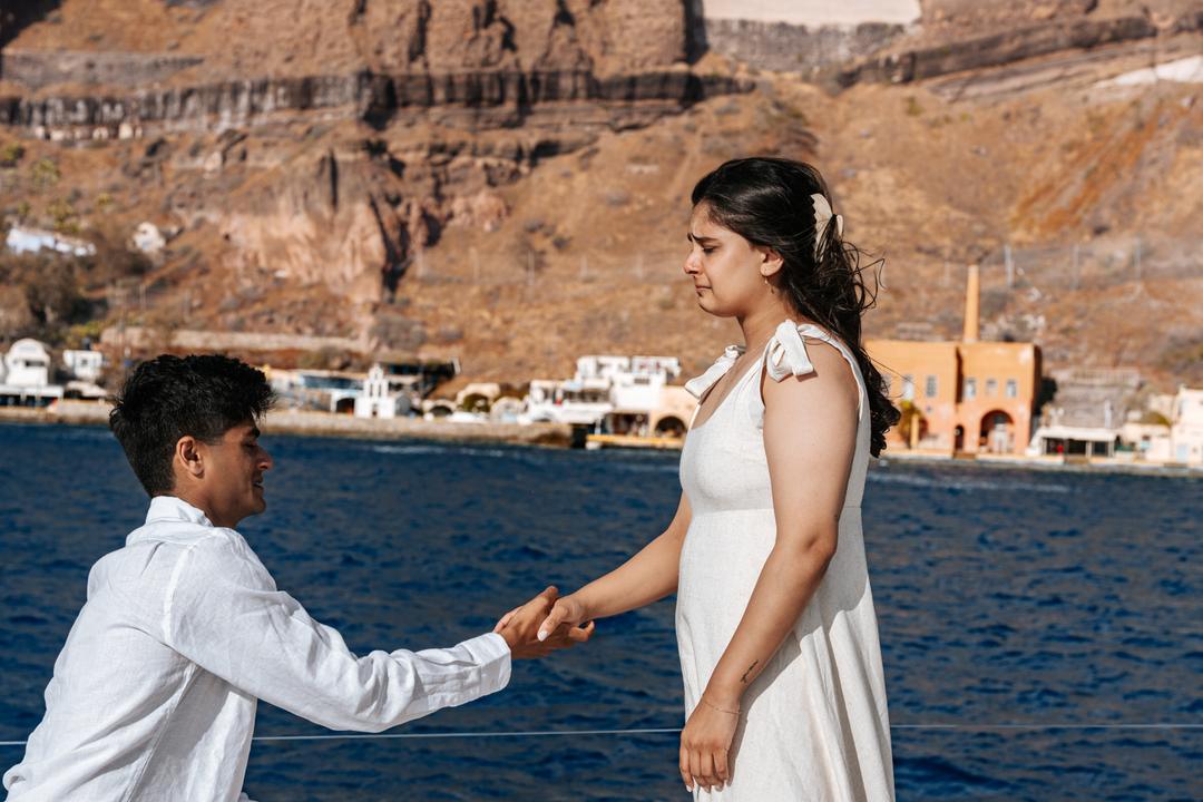 A man kneeling on deck reaching for her hand, a small harbor and the red cliffs of the caldera behind them