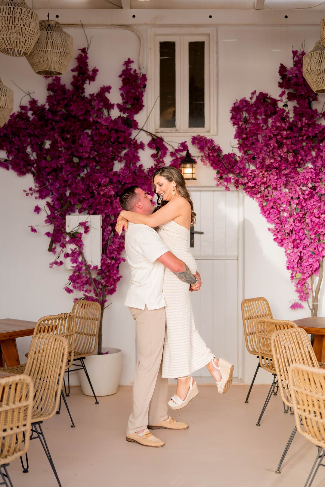 A man lifting a woman against a white wall covered in magenta bougainvillea, rattan chairs around them