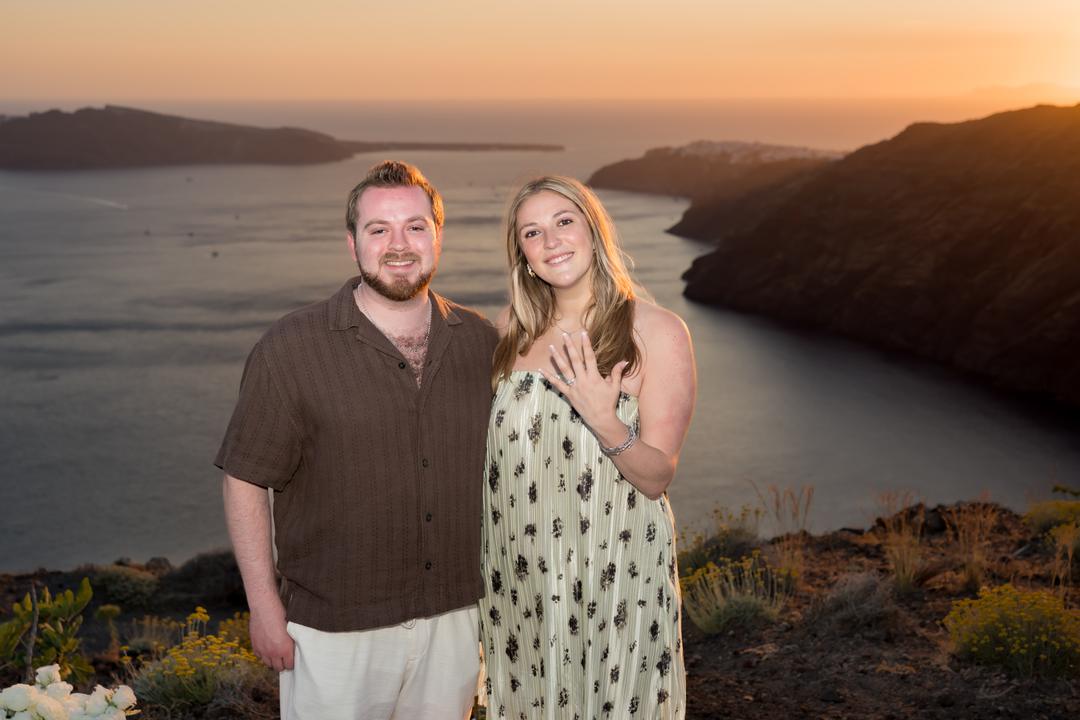 A couple standing together on the rim at sunset, her hand at her chest showing a new ring, the caldera falling away behind them