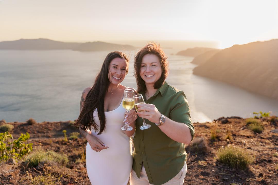 Two people touching champagne glasses on the rim path, the caldera and the far rim in soft backlight behind them
