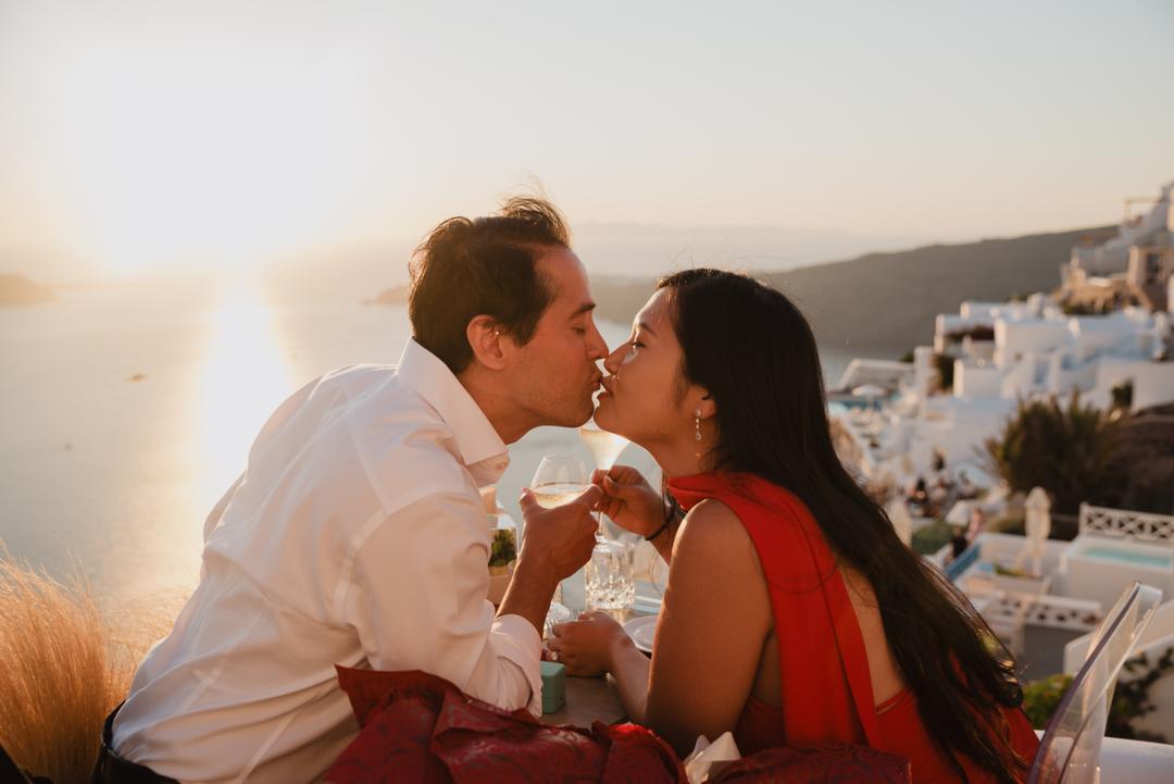 A couple kissing over champagne glasses at a terrace table, the white village and the sun low on the water behind them