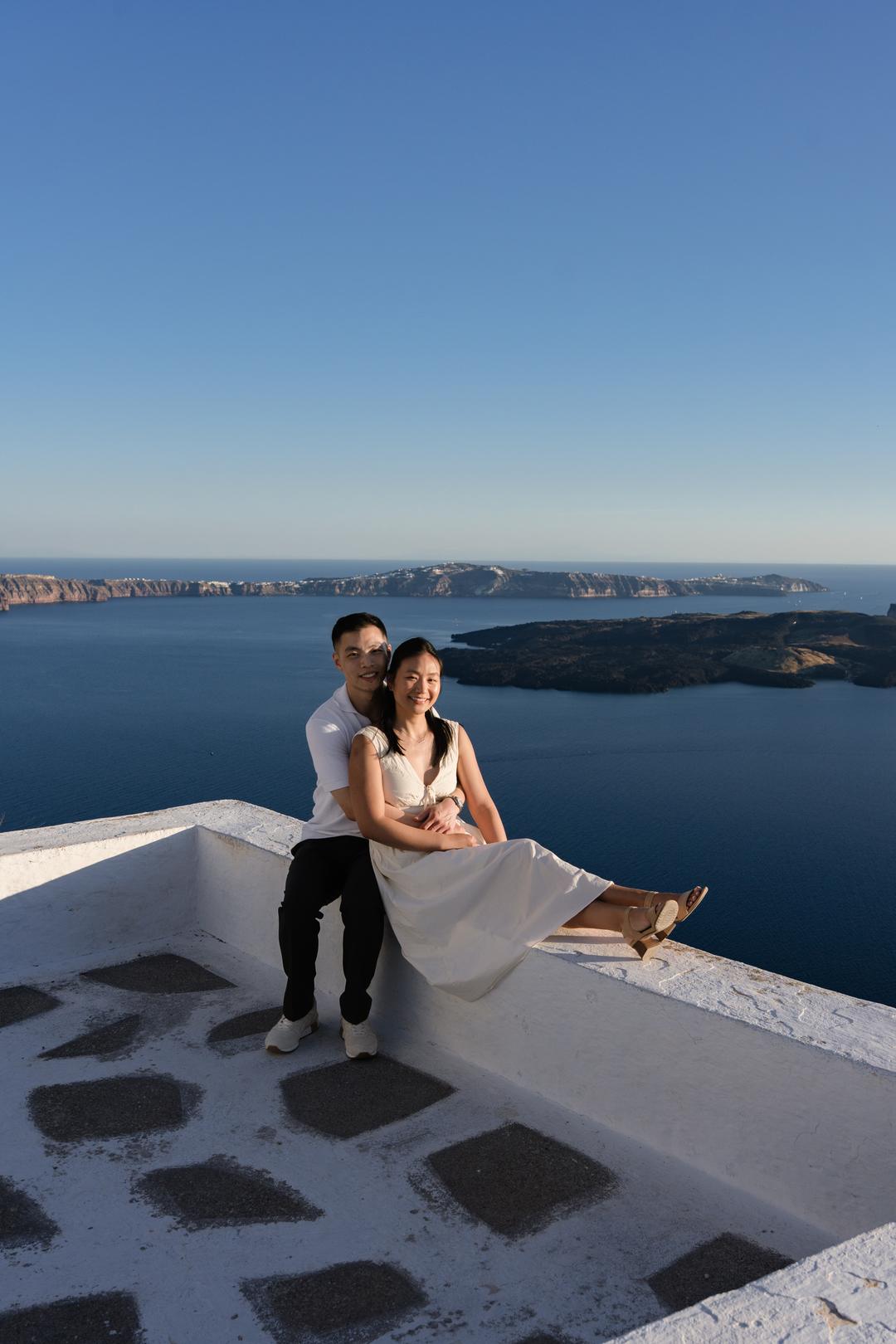 A couple sitting on the low wall of a whitewashed terrace, the caldera rim and the sea filling the frame behind them