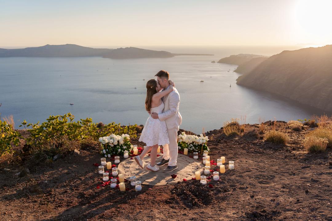 A couple embracing inside a wide circle of candles and white flowers on the ridge, the caldera and the far rim behind them at golden hour