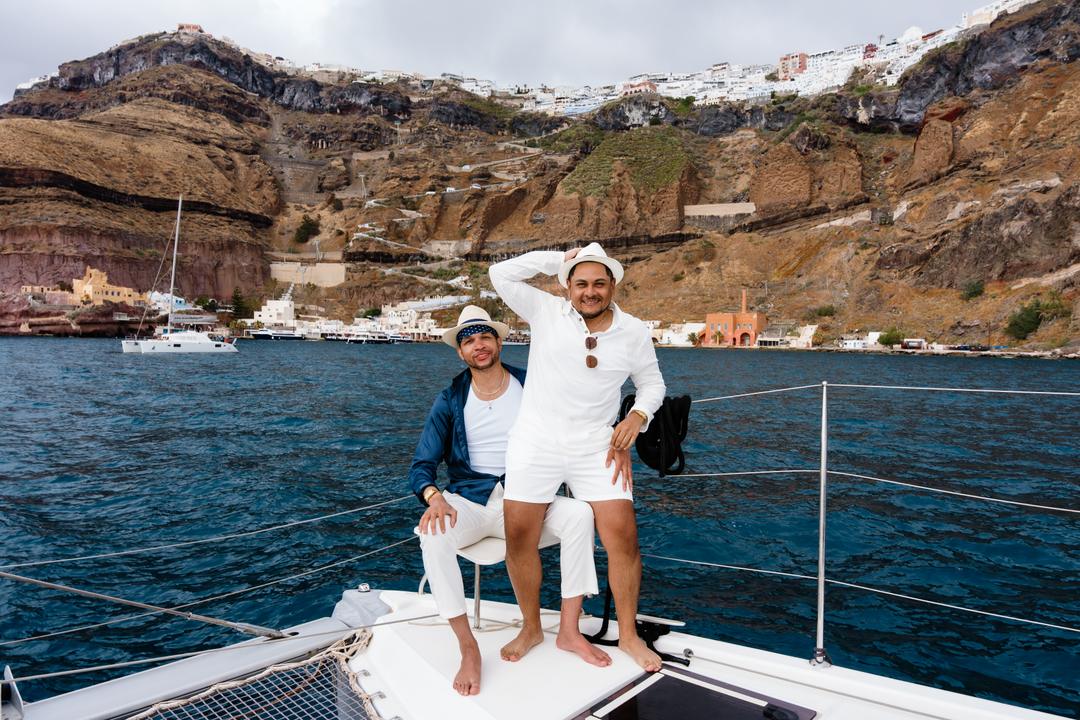One man seated and one standing at the bow of a catamaran, the caldera wall and the village above them