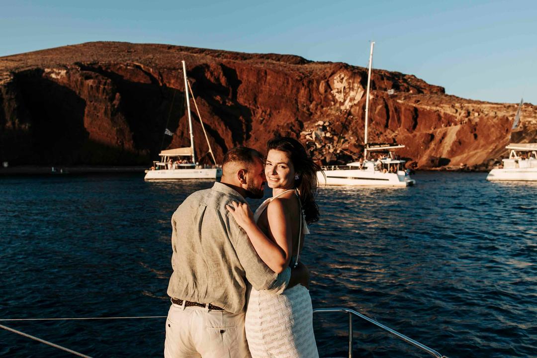 A couple embracing on deck in golden light, moored catamarans and the red volcanic cliffs of the caldera behind them