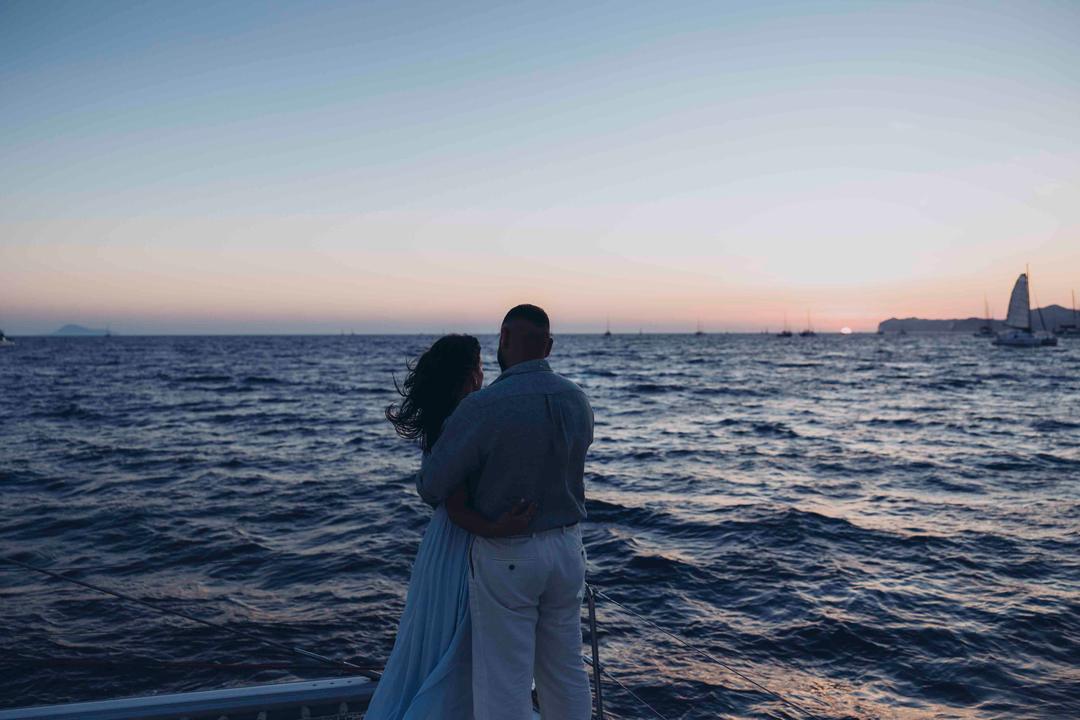 A couple at the rail with their backs to the camera, watching the sun go down over open water