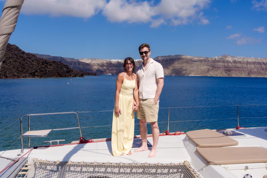 A couple standing on the netted foredeck of a catamaran over bright blue water, the caldera wall behind