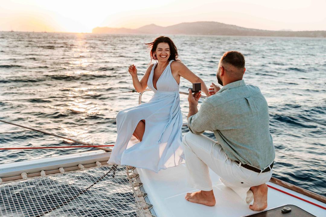 A woman laughing with her hands raised as a man kneels on the deck at sunset, the sun low on the water behind