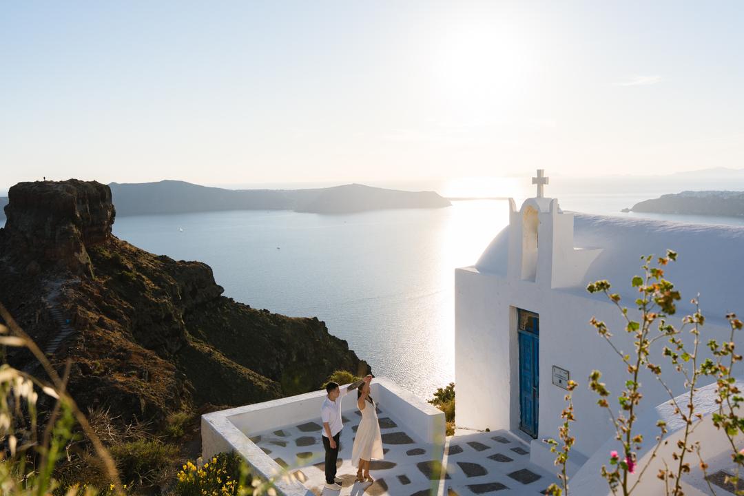 A couple on a small white terrace beside a chapel with a blue door, Skaros rock at one side and the low sun on the water