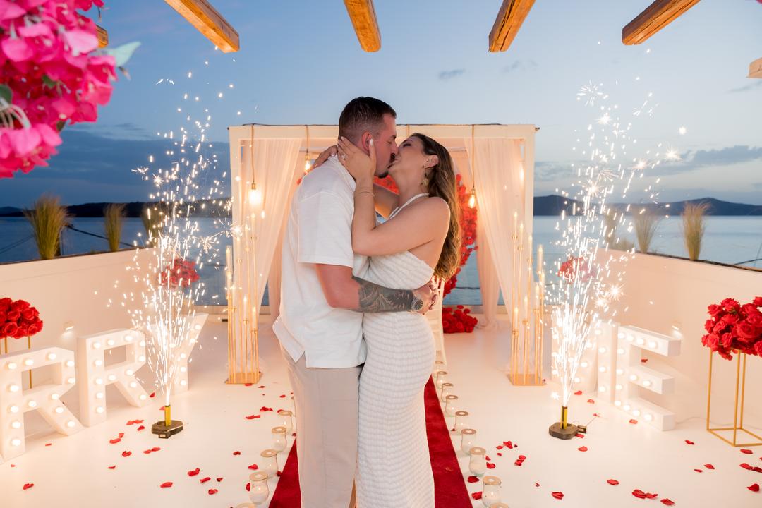 A couple kissing under a white arch while four cold-spark fountains throw gold light up around them at dusk