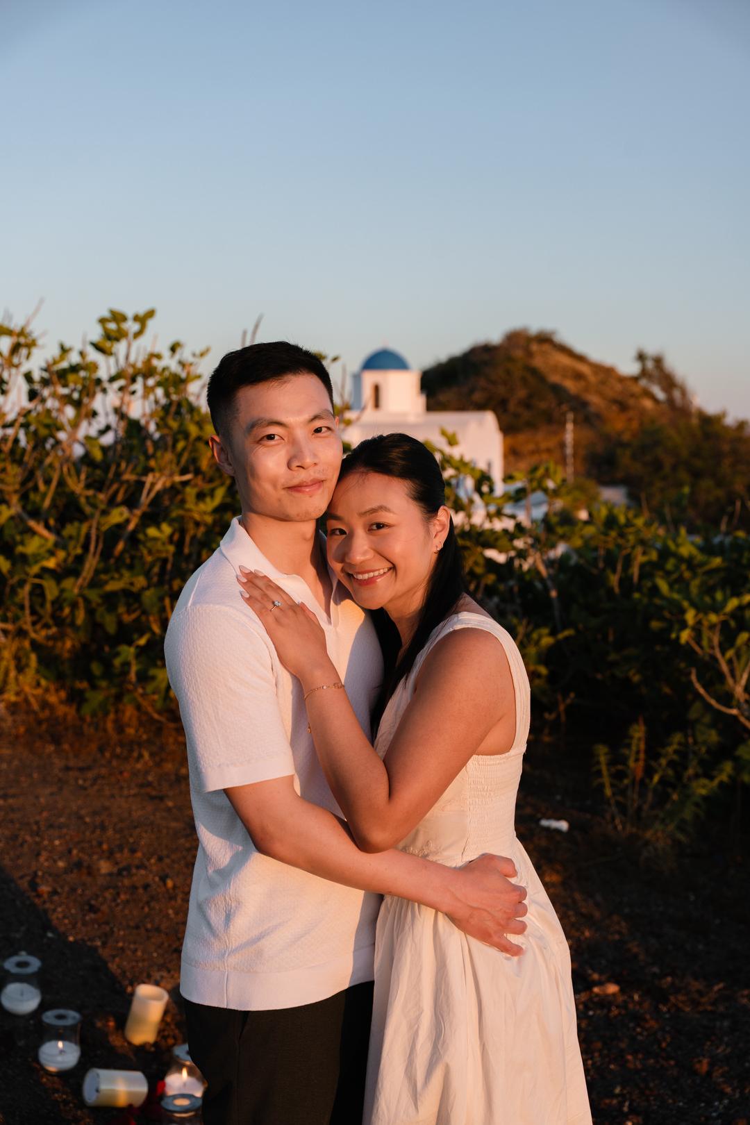 A couple holding each other in warm evening light with a blue-domed chapel on the ridge behind them and candles set out at their feet