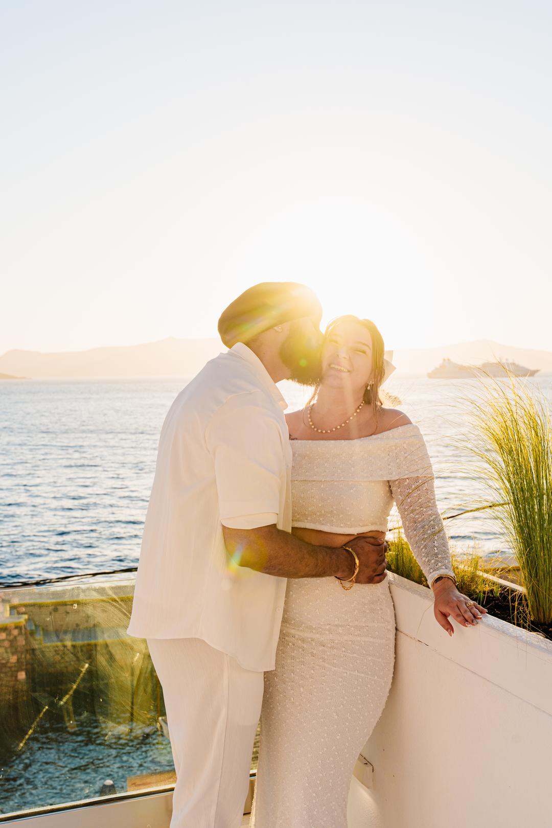 A couple at a glass terrace rail in low golden light, the sun breaking behind their shoulders over the sea