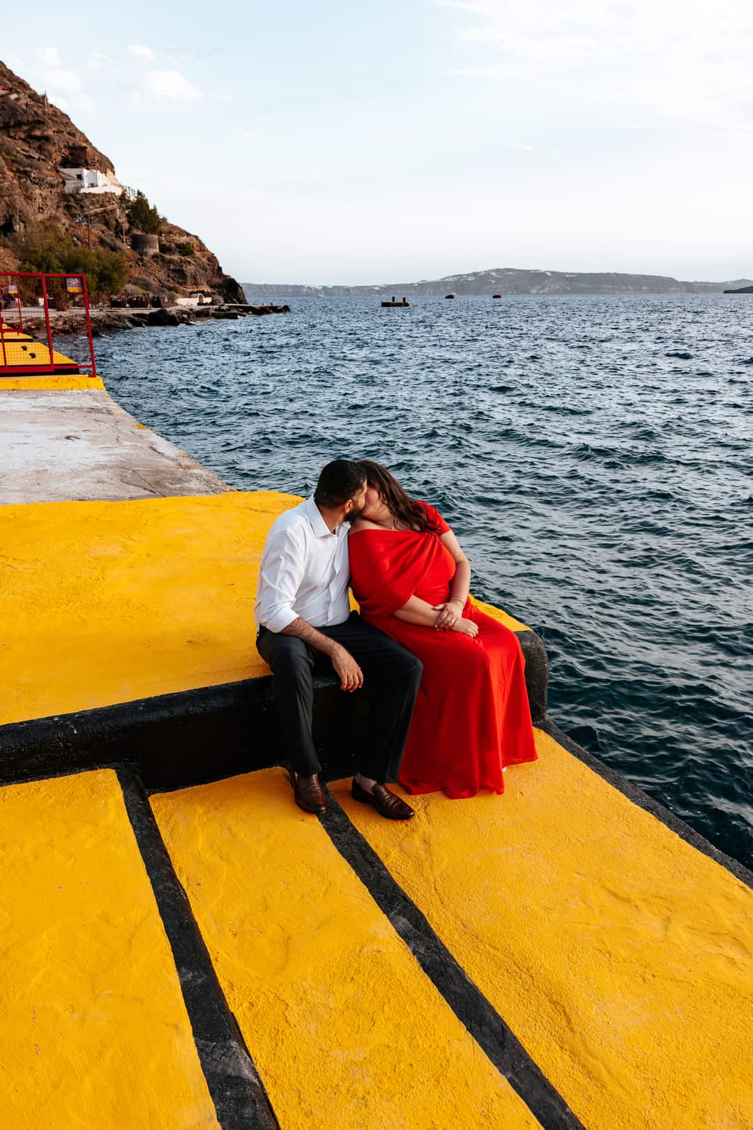 A couple sitting on a painted yellow jetty above dark blue water, the cliff and a small harbor behind