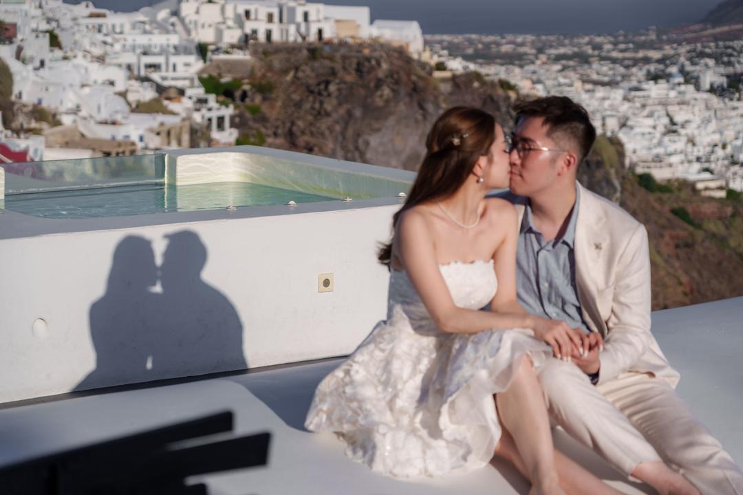 A couple sitting on the edge of an infinity pool terrace, their shadow on the white wall and a village stacked up the cliff behind