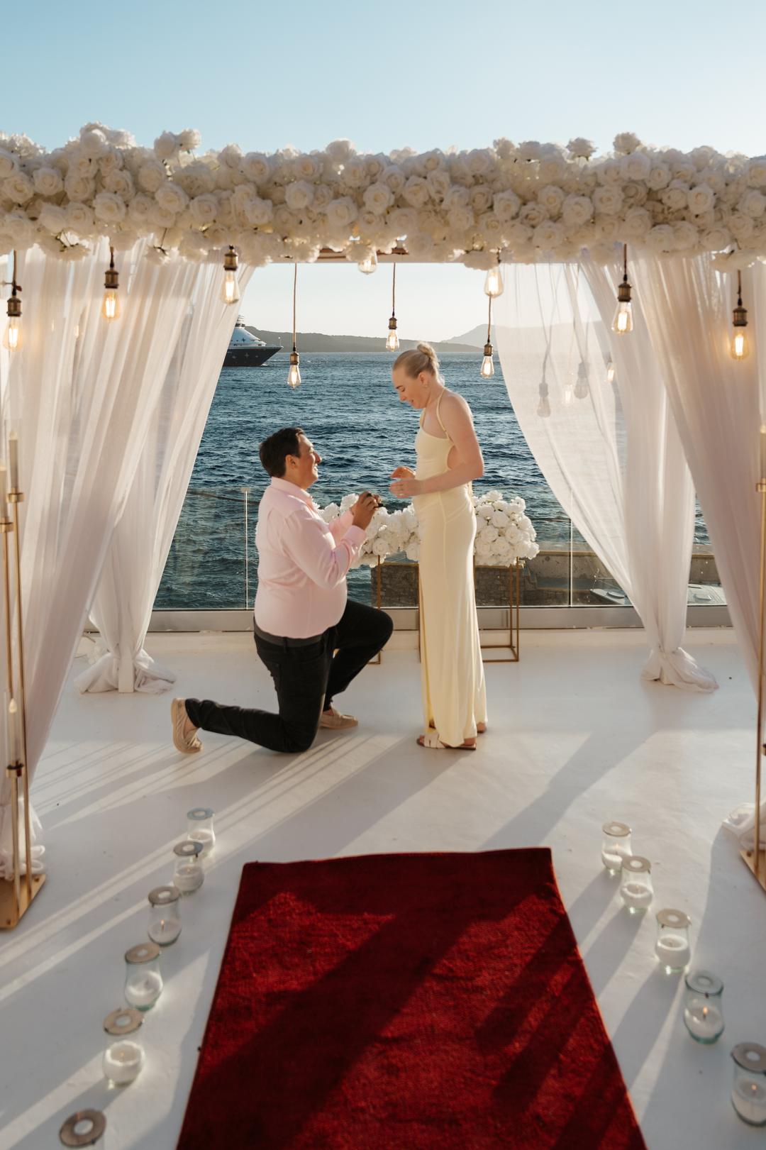 A man on one knee between white drapes at the end of a jetty, the open sea directly behind them