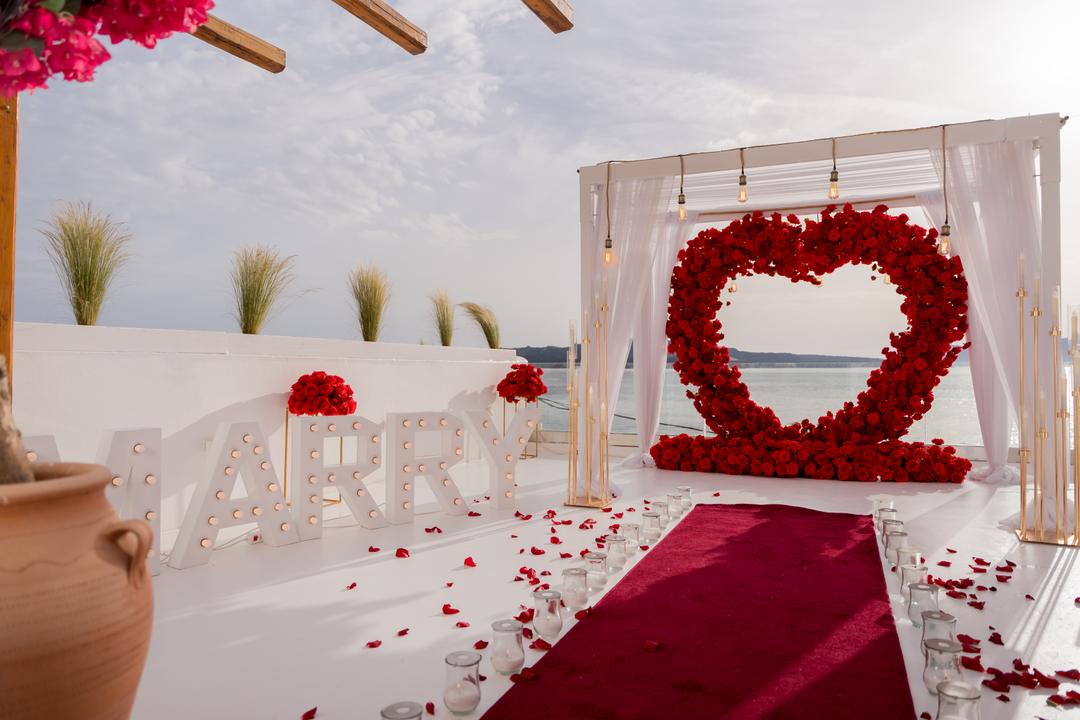 Lit MARRY ME letters in the foreground of a white terrace, a red rose heart arch and a red carpet beyond them