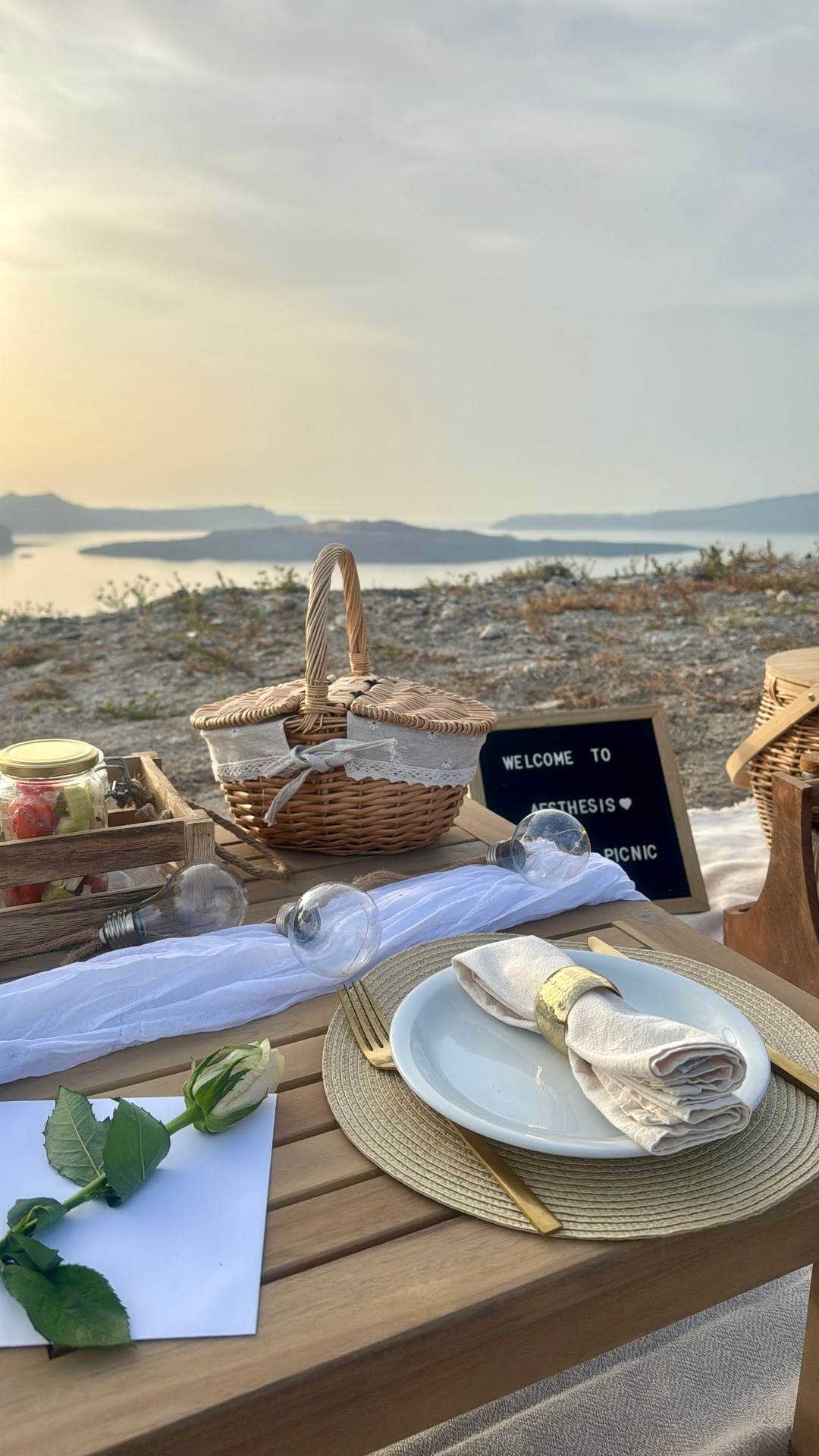 A low picnic table laid with plates, a wicker basket, preserve jars and a welcome board, set out on the rim above the caldera at sunset