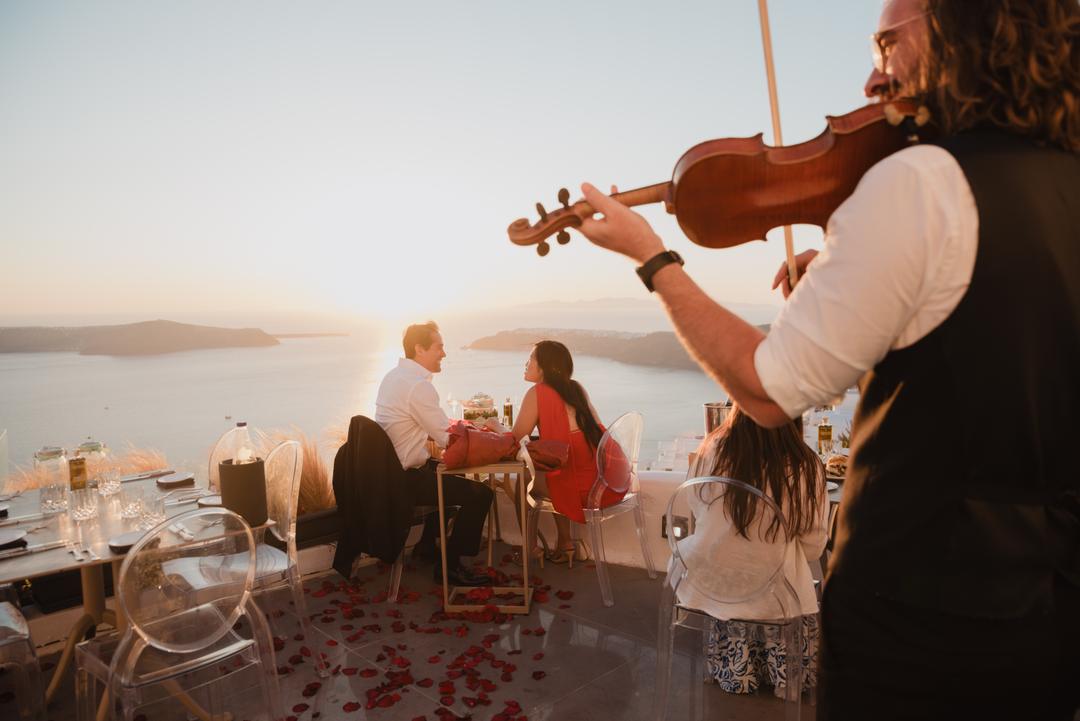 A violinist in the foreground with a couple seated at a laid table on the terrace behind, rose petals across the floor and the volcano beyond