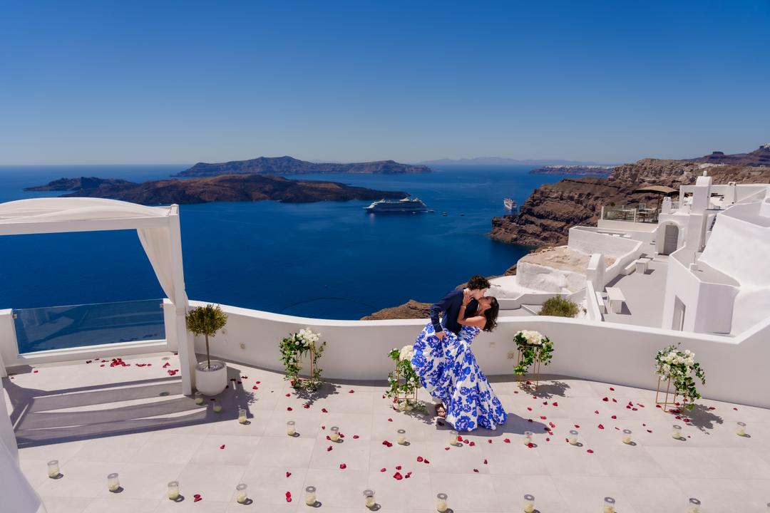A white terrace seen from above, a couple kissing at its far edge with the caldera, the volcano and a cruise ship beyond